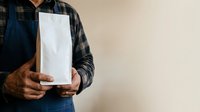 Worker in blue apron and plaid shirt holding blank white paper bag, studio shot with neutral background for retail and delivery mockups.