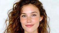 Young Caucasian woman with natural wavy brown hair and glowing skin smiling warmly at camera against white background, minimal makeup look emphasizing natural beauty.