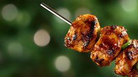 Grilled chicken skewers with caramelized glaze on metal stick against blurred green background. Barbecue meat kebabs with golden brown marinade for summer cooking.