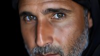 Close up portrait of mature Middle Eastern man with gray beard and intense gaze against dark background, dramatic lighting emphasizes facial features and expression.