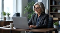 Mature woman with gray hair and glasses wearing dark blazer working on laptop at wooden desk in modern office with plants and natural light.
