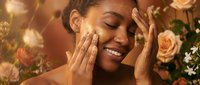 Young African American woman applying skincare cream with gentle massage motions surrounded by blooming roses in warm golden light for beauty routine.