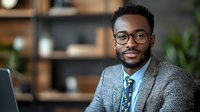 Young African American businessman wearing glasses and blue tie smiles confidently in modern office setting. Professional corporate portrait showing success and leadership.