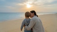 Mother and young son enjoying peaceful sunset moment together on sandy beach with calm ocean waves and golden sky creating intimate family bonding scene.