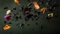 Scattered black tea leaves with vibrant rose petals and marigold flowers floating against dark green background, dramatic food photography composition.