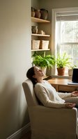 Young woman relaxing in comfortable armchair at home office with plants and wooden shelves creating peaceful workspace atmosphere for wellness and productivity.