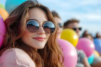 Young Caucasian woman with round sunglasses smiling at outdoor celebration with colorful balloons in background, expressing joy and happiness on sunny summer day.