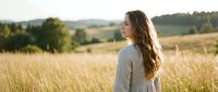 Young woman with long wavy hair in gray dress standing in golden wheat field during sunset, peaceful countryside landscape with rolling hills in background.