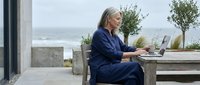 Senior businesswoman working remotely on laptop at modern outdoor terrace with ocean view, representing flexible work lifestyle and digital nomad concept.
