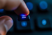 Finger pressing illuminated enter key on keyboard with blue backlight glow in dark environment, creating atmospheric technology mood for business concepts.