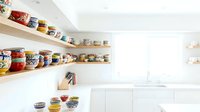 Bright modern white kitchen with open wooden shelves displaying colorful ceramic bowls collection, minimalist interior design with natural light.