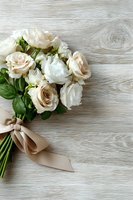 Elegant bouquet of cream roses and white peonies with green foliage tied with beige ribbon on rustic wooden background, perfect for wedding or celebration.