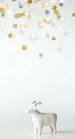Isolated white ceramic goat figurine standing against neutral background with scattered gold, silver, and cream confetti circles above, suitable for greeting cards, invitations, and festive design projects.