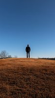 Silhouette of young man standing alone on hilltop against clear blue sky, wearing casual clothes, surrounded by autumn dry grass landscape at sunset.