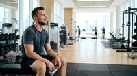 Caucasian male athlete in dark t-shirt and shorts resting on bench in modern gym facility with equipment and windows, fitness training break.