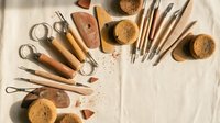 Clay sculpting and pottery tools arranged on linen fabric, including wooden-handled carving tools, sponges, and loop tools for ceramic art.