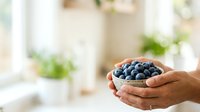 Young adult with medium skin tone holding a wooden bowl filled with fresh blueberries in bright natural light, suitable for food, nutrition, health and wellness marketing materials.