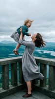 Mother lifting toddler son in air on mountain viewpoint balcony with scenic landscape background for family travel and bonding moments.