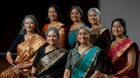 Group of elegant mature South Asian women wearing traditional silk saris in vibrant colors and ethnic jewelry posing together for formal portrait against dark background.