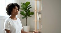 Young African American woman in white t-shirt sitting peacefully in modern minimalist home interior with green plant and wooden shelving unit in background.