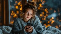 Young woman in cozy sweater using smartphone in bed with warm bokeh lights creating intimate evening atmosphere for relaxation and communication.