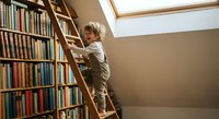 Young boy climbing wooden ladder in home library with floor-to-ceiling bookshelves under skylight for educational and learning concepts.