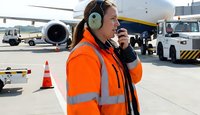 Female airport ground crew worker in orange safety vest using radio communication equipment on busy tarmac with aircraft and service vehicles in background.