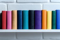 Colorful book spines arranged in rainbow order on white shelf against brick wall background, creating minimalist composition for education and reading concepts.