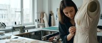 Young female fashion designer working on wedding dress in bright studio workspace examining lace details and fabric quality for bridal gown creation.