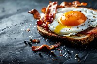 Crispy bacon strips and sunny side up egg on toasted rustic bread, seasoned with black pepper and salt on dark stone background. Runny yolk breakfast.