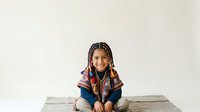 Young girl with braided hair and beaded braids wearing colorful striped shirt, sitting cross-legged on wooden surface against white background. Isolated portrait for design use.