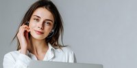 Young woman with gentle smile touching her face while working on laptop against gray background, portraying confidence and professionalism in business setting.
