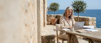 Woman with blonde hair working on laptop at outdoor desk overlooking Mediterranean sea, remote work and freelance lifestyle concept.