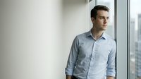 Young Caucasian businessman in light blue shirt standing by office window looking thoughtful and contemplative in modern corporate workspace.
