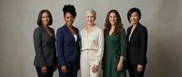 Diverse group of five professional women standing together in business attire against neutral background, representing corporate teamwork and leadership.