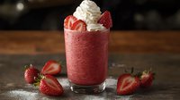 Fresh strawberry smoothie topped with whipped cream and sliced berries in glass on rustic wooden table, dark moody background with scattered ingredients.
