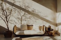 Minimalist living room interior with curved white sofa, potted tree, and dramatic shadow play on beige wall creating serene atmosphere for modern home decor.