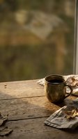 Rustic metal mug filled with dark coffee sits on weathered wooden table with autumn leaves and linen cloth, warm natural light streaming through window for cozy fall beverage styling.