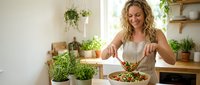 Happy blonde woman preparing fresh healthy salad in modern kitchen with herbs and plants for nutritious home cooking lifestyle.