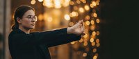 Young Caucasian woman in glasses and black outfit practicing martial arts outdoors with golden bokeh lights in the background.