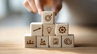 Hand arranging wooden blocks with business icons in pyramid formation, representing strategy planning and organizational hierarchy on wooden desk.