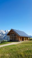 Modern wooden house with solar panels on roof in alpine mountain valley with green meadow and snow-capped peaks under clear blue sky, showcasing sustainable energy and eco-friendly architecture.