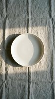 Empty white ceramic bowl on textured linen fabric with natural sunlight shadows creating minimalist kitchen tableware composition for cooking and dining concepts.