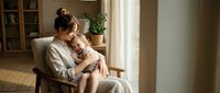 Young mother in white robe holding sleeping baby in cozy living room chair by window. Tender maternal bonding moment in warm natural light home interior.