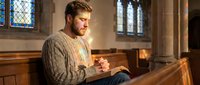 Young bearded man praying peacefully in historic church pew with warm sunlight streaming through stained glass windows creating spiritual atmosphere.