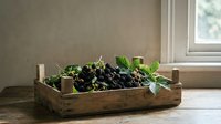 Black grapes with green leaves in rustic wooden crate by window, fresh produce display for kitchen decor and food styling.