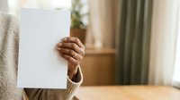 Person with medium skin tone holding blank white card or paper indoors, suitable for advertising, marketing campaigns, and promotional messaging.