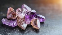 Raw amethyst and rose quartz crystals scattered on dark stone surface with soft natural lighting, showcasing mineral texture and transparency for wellness and spiritual concepts.