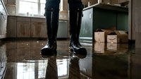Person in black rubber boots standing on flooded kitchen floor with water damage and cardboard boxes, home insurance claim documentation concept.