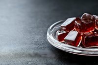 Red translucent jelly cubes on crystal glass plate against dark concrete background, selective focus with copy space for text or design elements.
